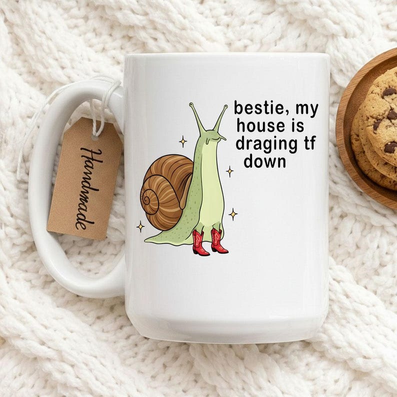 Op de afbeelding: Witte keramische mok met een cartoon slak met rode cowboylaarzen. De slak heeft een bruin huisje en is omringd door sterren. De tekst op de mok luidt "bestie, my house is dragging tf down". Een "Handmade" label hangt aan het handvat.