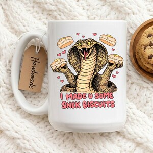 May include: White ceramic mug with a humorous illustration of a cobra holding biscuits. The cobra has a pink tongue and is surrounded by pink hearts and biscuits. Text on the mug reads "I MADE U SOME SNEK BISCUITS". A "Handmade" tag hangs from the handle.