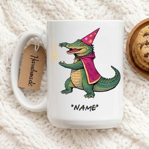 May include: White ceramic mug featuring a cartoon alligator wizard with a red cape and hat, conjuring golden sparkles. The mug has the text "*NAME*" and a "Handmade" tag.