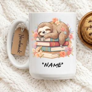 Puede incluir: Taza de cerámica blanca con una ilustración de un perezoso durmiendo sobre una pila de libros, adornada con flores. La taza tiene un asa grande y una etiqueta que dice "Handmade". El texto "*NAME*" está debajo del perezoso. Un plato de madera con galletas está en el fondo.