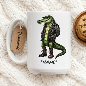 Könnte beinhalten: Weiße Keramik-Tasse mit einem Cartoon-Alligator, der eine schwarze Lederjacke, Sonnenbrille und Stiefel trägt. Der Alligator ist grün mit einem gelben Hemd. Auf der Tasse steht das Wort "*NAME*". Ein braunes Etikett mit dem Wort "Handmade" ist angebracht.