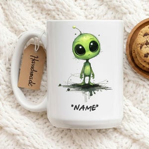 Op de afbeelding: Witte keramische mok met een cartoon alien ontwerp. De alien is groen met grote zwarte ogen en staat op een donkere spetter. De tekst "*NAME*" staat eronder. Een bruin label met "Handmade" hangt aan het handvat.