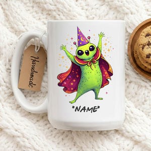 Op de afbeelding: Witte keramische mok met een groen alien-personage met een tovenaars hoed en cape. De alien heeft grote zwarte ogen en is omringd door sterren. De tekst "*NAME*" staat eronder. Een bruin label met "Handmade" is bevestigd.