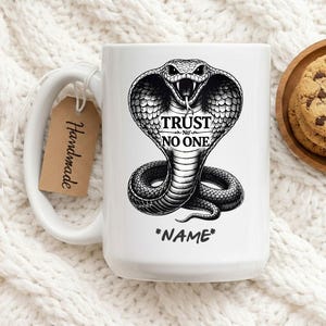 Puede incluir: Taza de cerámica blanca con una ilustración de cobra en blanco y negro. La taza tiene las palabras "TRUST NO ONE" y "*NAME*". Una pequeña etiqueta marrón con "Handmade" está unida al asa.