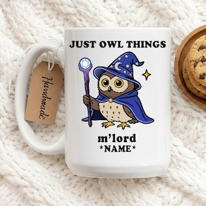 May include: White ceramic mug featuring a cartoon owl dressed as a wizard, holding a staff. The owl wears a blue cape and hat with stars. Text on the mug reads "JUST OWL THINGS" and "m'lord *NAME*". A small tag that says "Handmade" is attached.