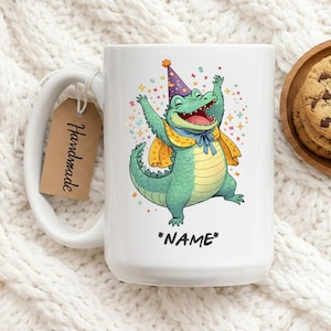 Peut inclure: Mug en céramique blanche avec un alligator de dessin animé portant un chapeau de fête et une écharpe, avec le texte "*NAME*". L'alligator est vert avec une écharpe jaune et bleue. Une étiquette "Handmade" est accrochée à la poignée. Des biscuits sont sur une assiette en bois.