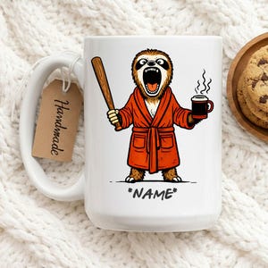May include: White ceramic mug featuring a cartoon sloth in an orange robe, holding a baseball bat and a coffee mug. The sloth has an open mouth and the text "*NAME*" is below. A "Handmade" tag hangs from the handle.
