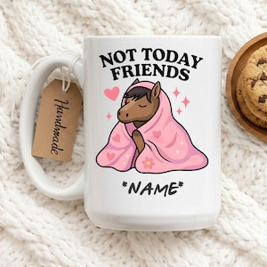 Pode incluir: Caneca de cerâmica branca com um design divertido. A caneca apresenta um cavalo de desenho animado envolto em um cobertor rosa com corações e uma flor. O texto na caneca diz "NOT TODAY FRIENDS" e "*NAME*". Uma etiqueta feita à mão está anexada.