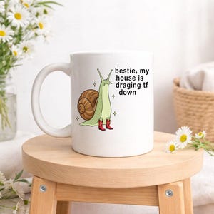 Op de afbeelding: Witte keramische mok met een cartoon slak ontwerp. De slak heeft een bruin huis, een groen lichaam en rode laarzen. De tekst op de mok luidt: "bestie, my house is dragging tf down."
