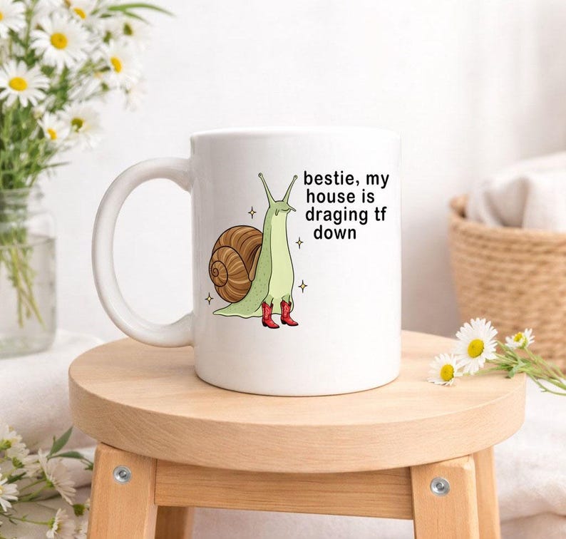 Op de afbeelding: Witte keramische mok met een cartoon slak ontwerp. De slak heeft een bruin huis, een groen lichaam en rode laarzen. De tekst op de mok luidt: "bestie, my house is dragging tf down."
