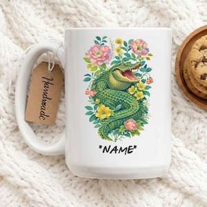 Puede incluir: Taza de cerámica blanca con un diseño de caimán verde rodeado de flores rosas y amarillas. La taza tiene un asa grande y una etiqueta que dice "Handmade". El texto "*NAME*" está debajo del caimán.