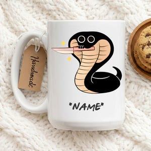 Könnte beinhalten: Weiße Keramiktasse mit einem Cartoon-Kobra-Design, die ein Messer hält, und dem Text "*NAME*". Ein braunes Etikett mit "Handmade" ist angebracht. Kekse liegen auf einem Holztablett.