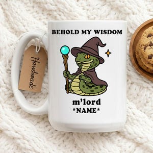 Könnte beinhalten: Weiße Keramik-Tasse mit einem Cartoon-Schlangen-Zauberer mit braunem Hut und Umhang, der einen Stab mit einer blauen Kugel hält. Auf der Tasse steht "BEHOLD MY WISDOM" und "m'lord *NAME*". Ein "Handmade"-Etikett ist angebracht.