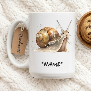 Könnte beinhalten: Weiße Keramiktasse mit einer Aquarellillustration einer Schnecke mit braunem Gehäuse und dem Text "*NAME*". Ein braunes Etikett mit der Aufschrift "Handmade" ist an der Tasse befestigt. Die Tasse steht auf einer weißen, strukturierten Oberfläche.