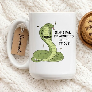 Puede incluir: Taza de cerámica blanca con un diseño de cobra de dibujos animados en verde y negro. El texto dice "Snake Pal, I'm about to strike TF out". Una etiqueta marrón con "Handmade" está adjunta. La taza está sobre una superficie blanca.