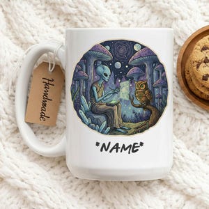 Może przedstawiać: Biały ceramiczny kubek z kolorową ilustracją kosmity, sowy i grzybów. Kosmita trzyma świecącą tabliczkę. Kubek ma napis "*NAME*" i metkę "Handmade".