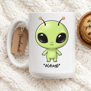 Peut inclure: Mug en céramique blanche avec un dessin d'extraterrestre de dessin animé. L'extraterrestre est vert avec de grands yeux noirs, des antennes oranges et un petit sourire. Le mot "*NAME*" est en dessous. Une étiquette marron avec "Handmade" est attachée.