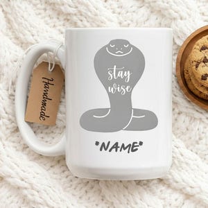 Puede incluir: Taza de cerámica blanca con una ilustración de cobra gris y las palabras "stay wise" y "*NAME*". Una etiqueta marrón con "Handmade" está adjunta a la taza. La taza está sobre una superficie de punto blanca.