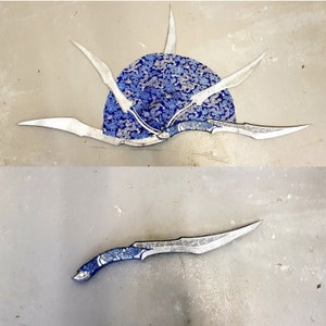 Puede incluir: Cinco cuchillos de lanzamiento plateados con mangos con patrones azules y plateados dispuestos en forma de estrella alrededor de un círculo con patrones azules y plateados.