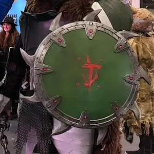 Puede incluir: Un escudo verde y plateado con un símbolo rojo de la cara de un demonio y un tridente. El escudo tiene púas alrededor del borde.