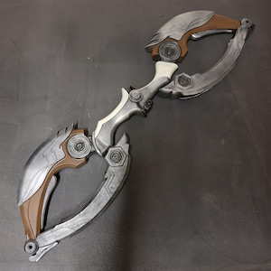 Könnte beinhalten: Eine silber-braune Requisitenwaffe mit gebogenem Klingendesign. Die Requisite weist komplizierte Details auf, darunter ein weißer Griff und dekorative Akzente. Die Waffe hat eine futuristische Ästhetik mit metallischem Finish und symmetrischem Design.