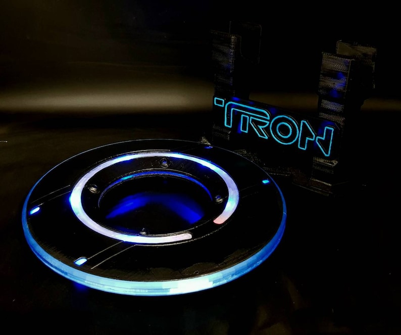 May include: A black and blue light-up disc with a glowing blue ring around the edge. The disc is sitting on a black surface next to a black stand with the word "TRON" in blue lights.