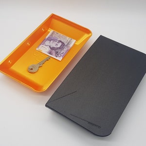 May include: Two small, rectangular trays, one orange and one black. The orange tray has a key and a British pound note inside.