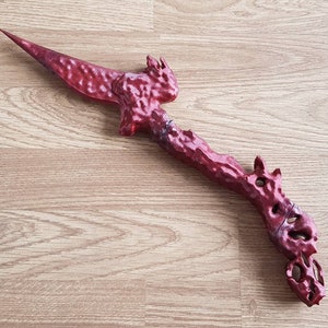 Może przedstawiać: Dekoracyjny miecz w kształcie smoka, czerwony, szczegółowy, z ostrym końcem. Miecz wykonany jest z fakturowanego materiału i ma ciemnoczerwony kolor.