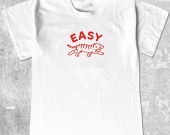 May include: White t-shirt with a red graphic of a tiger and the word "EASY" above it. The tiger is in a running pose, with stripes and details. The shirt has a crew neck and short sleeves.