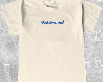 May include: Cream-colored t-shirt with the word "Overstimulated" in blue script across the chest. The shirt has a classic crew neck and short sleeves. The fabric appears to be a soft cotton blend.