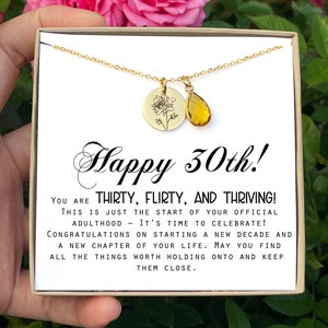 May include: A gold necklace with a small flower charm and a teardrop-shaped amber stone charm. The necklace is in a white box with the text "Happy 30th! You are thirty, flirty, and thriving! This is just the start of your official adulthood - it's time to celebrate! Congratulations on starting a new decade and a new chapter of your life. May you find all the things worth holding onto and keep them close."