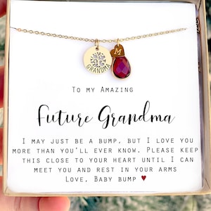 May include: A gold necklace with a charm that says "Grandma" and a red gemstone charm with the letter "M". The necklace is in a white box with the text "To my amazing Future Grandma I may just be a bump, but I love you more than you'll ever know. Please keep this close to your heart until I can meet you and rest in your arms Love, Baby Bump".
