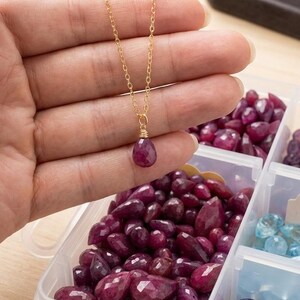 May include: A gold chain necklace featuring a single teardrop-shaped faceted dark pink gemstone pendant, secured with gold wire wrapping. The necklace is held above a clear plastic organizer box filled with various loose faceted gemstones.