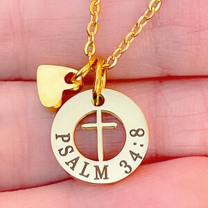 Op de afbeelding: Gouden ketting met een klein hartvormig bedeltje en een ronde hanger met een kruisuitsparing en de woorden "PSALM 34:8" gegraveerd rond de rand.