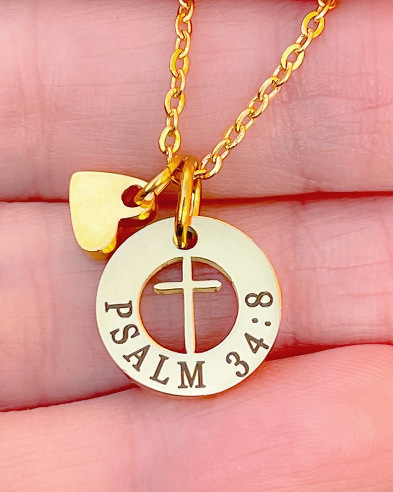 Op de afbeelding: Gouden ketting met een klein hartvormig bedeltje en een ronde hanger met een kruisuitsparing en de woorden "PSALM 34:8" gegraveerd rond de rand.