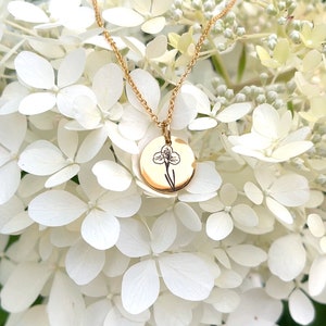 May include: Gold-colored necklace with a round pendant featuring a black floral design. The necklace hangs against a backdrop of white hydrangea flowers, creating a delicate and elegant aesthetic.