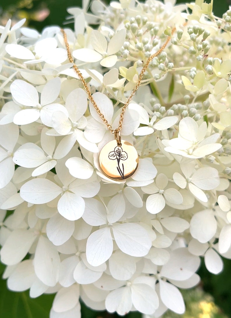 May include: Gold-colored necklace with a round pendant featuring a black floral design. The necklace hangs against a backdrop of white hydrangea flowers, creating a delicate and elegant aesthetic.