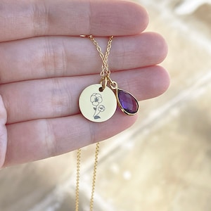 May include: A gold-colored necklace featuring a round pendant with a floral design and a teardrop-shaped purple gemstone. The delicate chain and pendants are held in a hand, showcasing the jewelry's details.