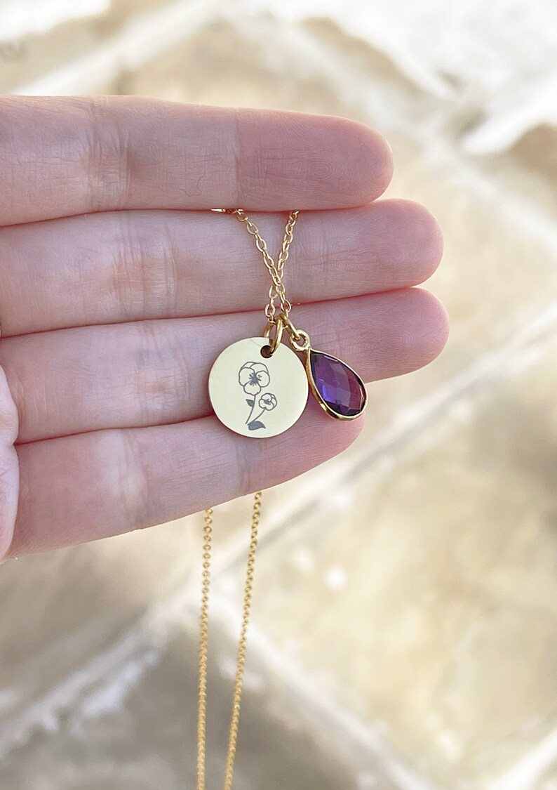 May include: A gold-colored necklace featuring a round pendant with a floral design and a teardrop-shaped purple gemstone. The delicate chain and pendants are held in a hand, showcasing the jewelry's details.