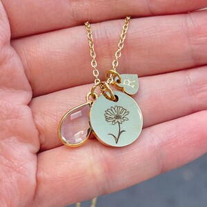 May include: Gold necklace with three charms: a clear teardrop-shaped crystal, a round charm with a delicate flower design, and a heart charm with the letter "R" engraved on it.
