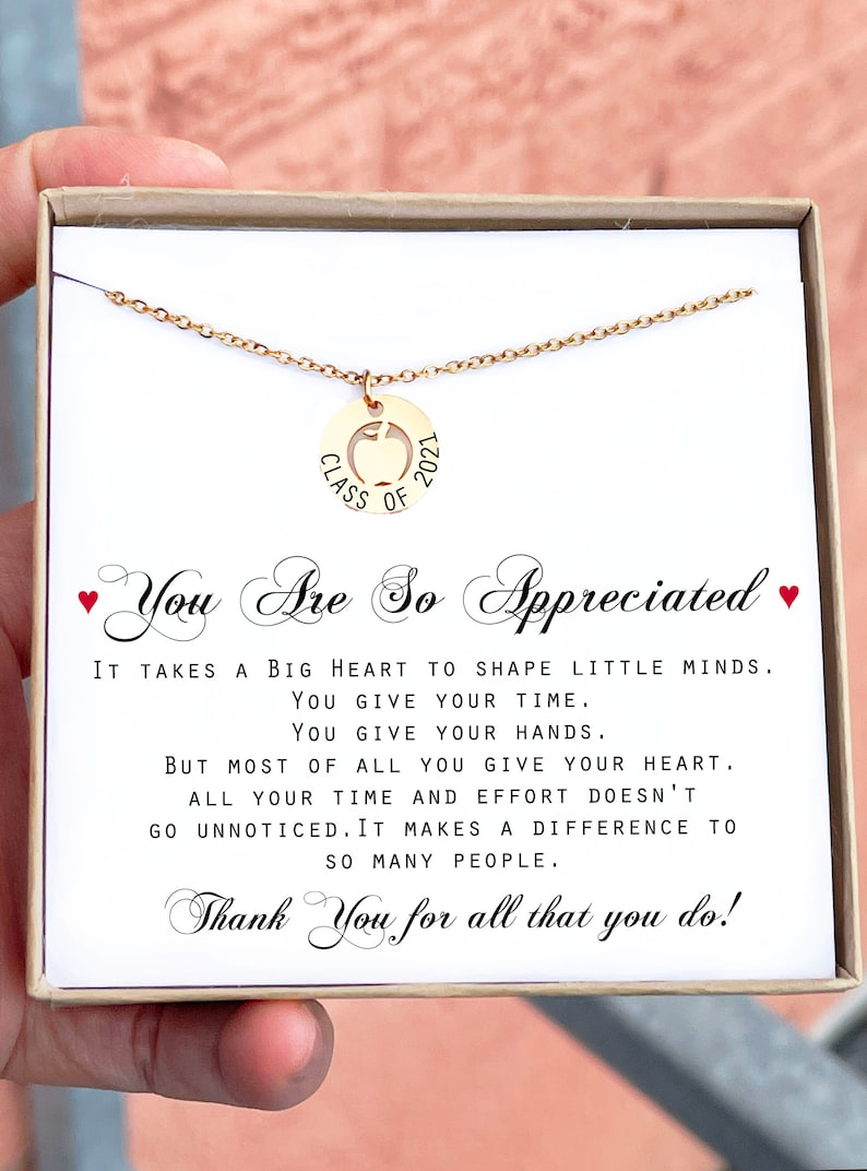 May include: A gold necklace with a pendant that says "Class of 2021" with an apple cut out in the center. The necklace is in a box with a message that reads "You are so appreciated. It takes a big heart to shape little minds. You give your time. You give your hands. But most of all you give your heart. All your time and effort doesn't go unnoticed. It makes a difference to so many people. Thank you for all that you do!"