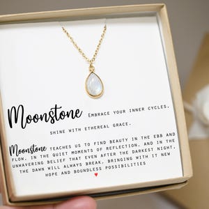 May include: A white moonstone necklace in a gift box with a card that reads "Moonstone Embrace your inner cycles. Shine with ethereal grace. Moonstone teaches us to find beauty in the ebb and flow. In the quiet moments of reflection, and in the unwavering belief that even after the darkest night, the dawn will always break, bringing with it new hope and boundless possibilities."