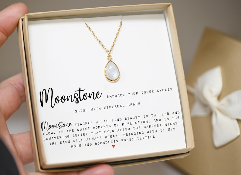 May include: A white moonstone necklace in a gift box with a card that reads "Moonstone Embrace your inner cycles. Shine with ethereal grace. Moonstone teaches us to find beauty in the ebb and flow. In the quiet moments of reflection, and in the unwavering belief that even after the darkest night, the dawn will always break, bringing with it new hope and boundless possibilities."