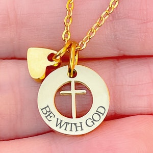 Op de afbeelding: Gouden ketting met een ronde hanger met een kruisuitsparing en de woorden "BE WITH GOD". Een kleine hartvormige hanger hangt boven de hanger.