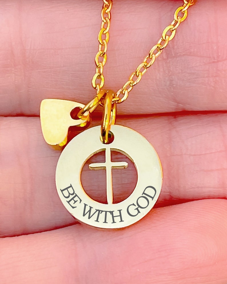 Op de afbeelding: Gouden ketting met een ronde hanger met een kruisuitsparing en de woorden "BE WITH GOD". Een kleine hartvormige hanger hangt boven de hanger.