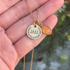 Op de afbeelding: Gouden ketting met een ronde hanger met de tekst "Gefeliciteerd 2022" en een kleine traanvormige hanger van oranje edelsteen.