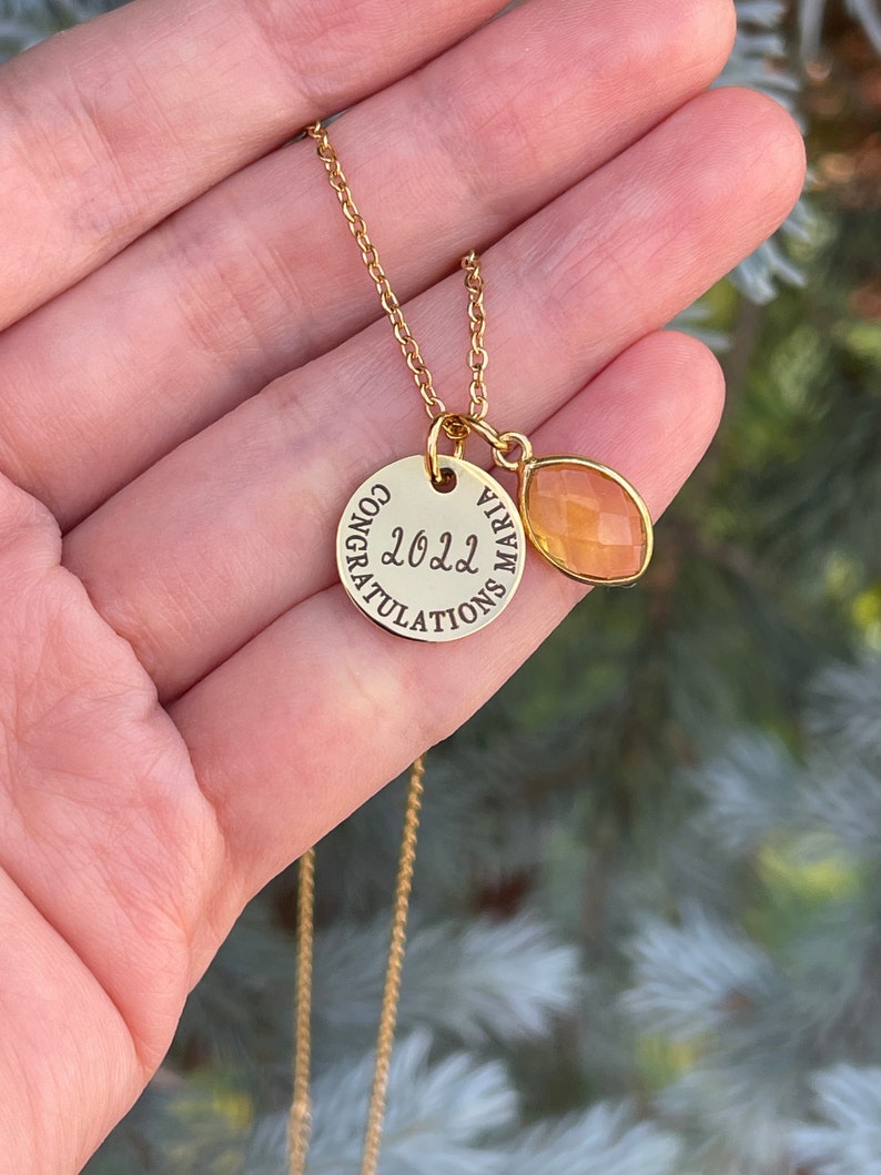 Op de afbeelding: Gouden ketting met een ronde hanger met de tekst "Gefeliciteerd 2022" en een kleine traanvormige hanger van oranje edelsteen.