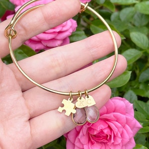 May include: Gold-colored bangle bracelet with three charms. Charms include a puzzle piece, a heart-shaped charm with the letter "K", and a teardrop-shaped charm with the letter "C". The bracelet is displayed in front of pink roses.