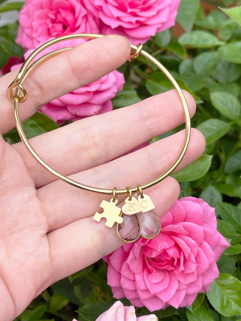 May include: Gold-colored bangle bracelet with three charms. Charms include a puzzle piece, a heart-shaped charm with the letter "K", and a teardrop-shaped charm with the letter "C". The bracelet is displayed in front of pink roses.