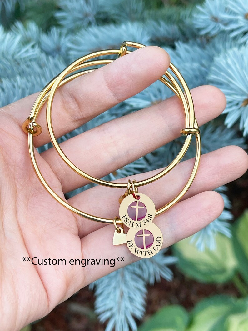 Op de afbeelding: Twee gouden armbandjes met een bedel met de tekst "Psalm 34:8 Wees met God" met een kruis en een hartbedel.